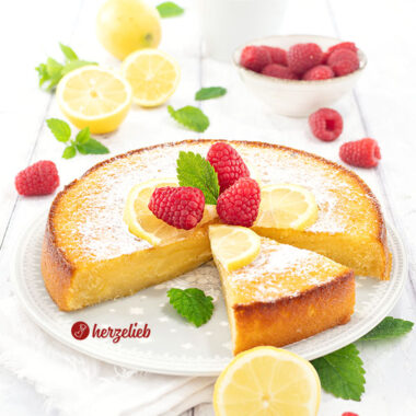 Frisch gebackener und angeschnittener schwedischer Zitronenkuchen nach einem Kladdkaka Rezept aus Schweden von herzelieb auf einem weißen Teller, dekoriert mit Zitronenscheiben, Himbeeren und Minzblättern.