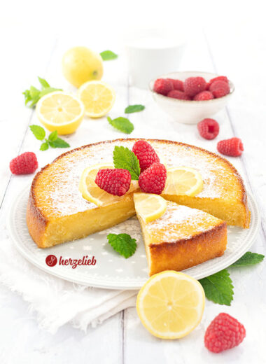 Frisch gebackener und angeschnittener schwedischer Zitronenkuchen nach einem Kladdkaka Rezept aus Schweden von herzelieb auf einem weißen Teller, dekoriert mit Zitronenscheiben, Himbeeren und Minzblättern.