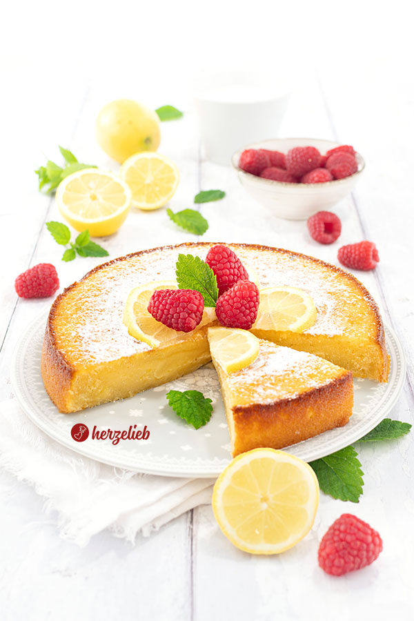 Frisch gebackener und angeschnittener schwedischer Zitronenkuchen nach einem Citronkladdka Rezept aus Schweden von herzelieb auf einem weißen Teller, dekoriert mit Zitronenscheiben, Himbeeren und Minzblättern.