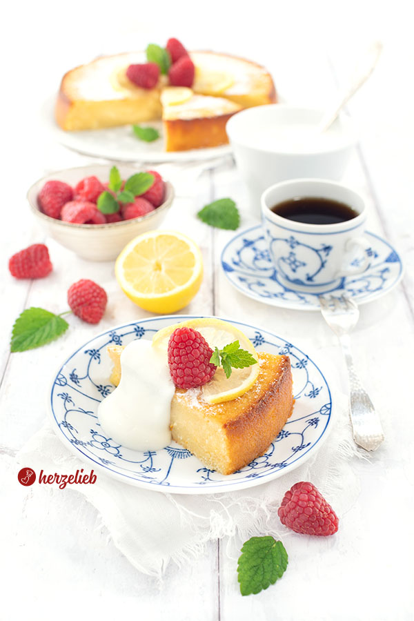 Ein Stück vom schwedischer Zitronenkuchen nach einem Citronkladdkaka Rezept aus Schweden von herzelieb, serviert auf einem weiß-blauen Teller mit einer Portion Sahne, Himbeeren. Im Hintergrund eine Tasse Kaffee, Früchte, ein Schälchen Sahne und der abgeschnittenen Kuchen.