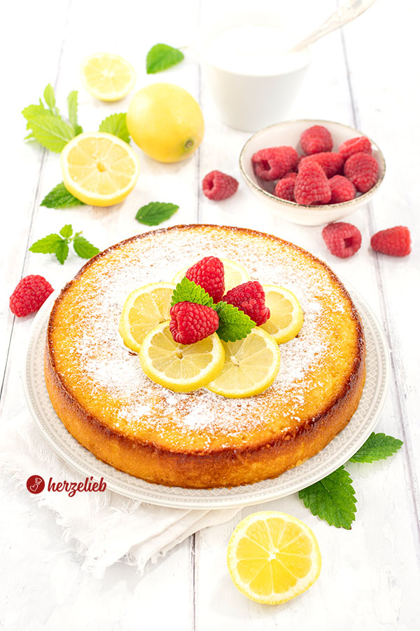 Ganzer schwedischer Zitronenkuchen nach einem Kladdkaka Rezept aus Schweden , bestreut mit Puderzucker und garniert mit Zitronenscheiben, Himbeeren sowie frischen Zitronenmelisseblättern. Hinter dem Citronkladdkaka Zitronen, Himbeeren in einer Schale und ein Schälchen Crème légère.