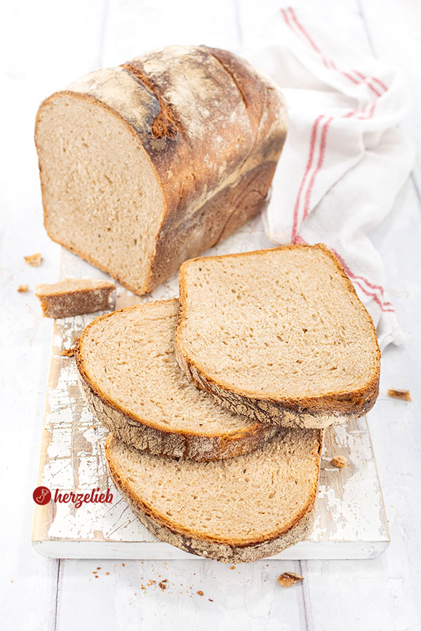 Frisch gebackenes Brot mit knuspriger Kruste nach einem Kefirbrot Rezept von herzelieb mit knuspriger Kruste, angeschnitten in mehrere Scheiben, auf einem weißen Holzbrett neben einem weißen Küchentuch mit roten Streifen.