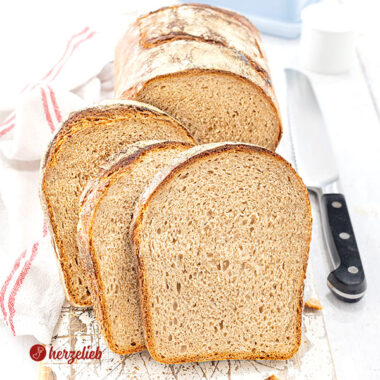 Knuspriges Brot mit Dinkel, Kefir und Roggen nach einem Kefirbrot Rezept von herzelieb, in mehrere Scheiben geschnitten, die vor dem angeschnittenem Brotlaib stehen. Alles ist auf einem weißen Holzbrett angerichtet, rechts daneben ein Messer und ein weißes Küchentuch mit roten Streifen.