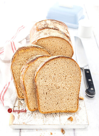 Knuspriges Brot mit Dinkel, Kefir und Roggen nach einem Kefirbrot Rezept von herzelieb, in mehrere Scheiben geschnitten, die vor dem angeschnittenem Brotlaib stehen. Alles ist auf einem weißen Holzbrett angerichtet, rechts daneben ein Messer und ein weißes Küchentuch mit roten Streifen.