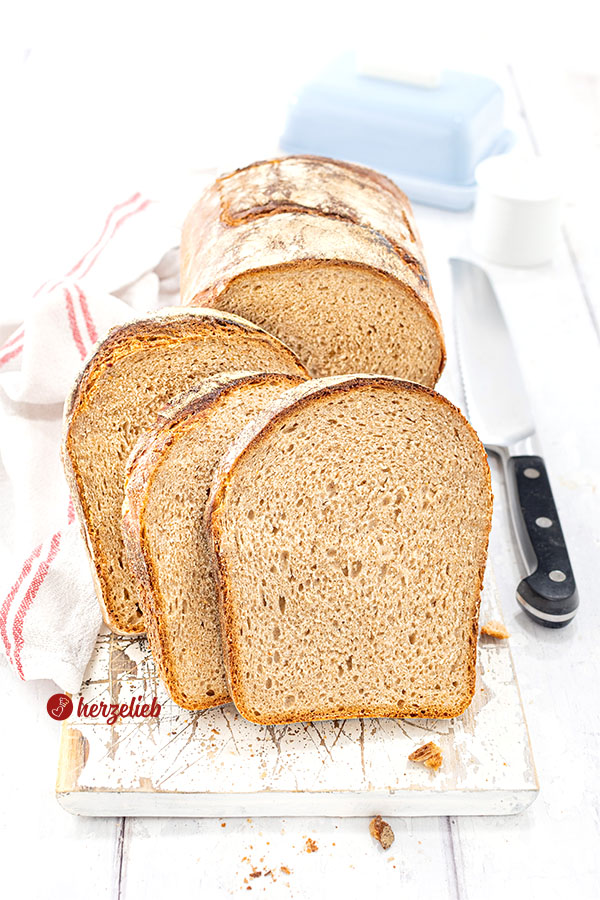 Knuspriges Brot mit Dinkel, Kefir und Roggen nach einem Kefirbrot Rezept von herzelieb, in mehrere Scheiben geschnitten, die vor dem angeschnittenem Brotlaib stehen. Alles ist auf einem weißen Holzbrett angerichtet, rechts daneben ein Messer und ein weißes Küchentuch mit roten Streifen.