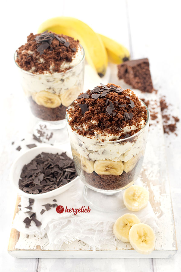 Nachtisch in Zwei Gläsern nach einem Maulwurfkuchen Dessert im Glas Rezept von herzelieb, geschichtet mit Schokokuchenbröseln, Bananenscheiben und Sahnecreme, garniert mit Schokoraspeln. Im Hintergrund liegen Bananen, neben den Gläsern eine Schale mit Schokostückchen und Bananenscheiben auf einem weißen Holzbrett.