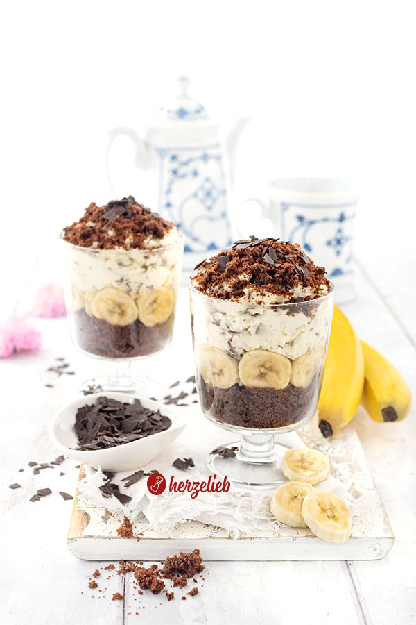 Zwei Gläser mit einem Schichtdessert nach einem Maulwurfkuchen Dessert im Glas Rezept, geschichtet aus Schokokuchen, Bananenscheiben, Sahnecreme und Schokoraspeln, daneben Bananen und Schokostückchen, im hellen Kaffeetafel-Setting.