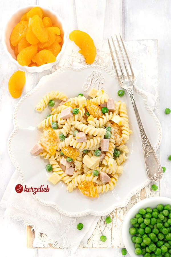 Frisch angerichteter Nudelsalat mit Mandarinen auf einem dekorativen weißen Teller, mit kleinen Schalen mit Erbsen und Mandarinenstücken.
