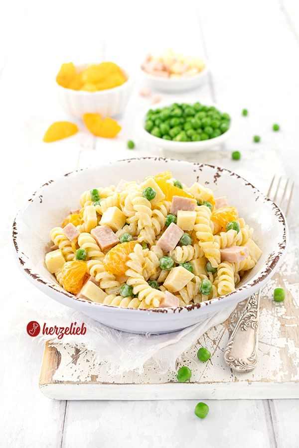 Eine rustikale, weiße Schale gefüllt mit Pastasalat nach einem Nudelsalat mit Mandarinen Rezept, umgeben von kleinen Schälchen mit grünen Erbsen, Mandarinenstücken und Fleischwurstwürfeln und Käsewürfeln
