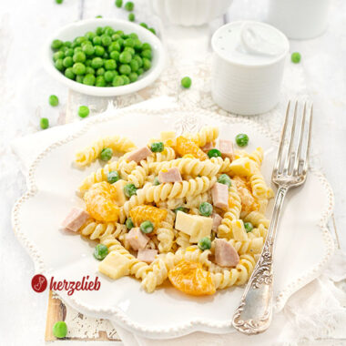 Nahaufnahme eines Pastasalats nach einem Nudelsalat mit Mandarinen Rezept von Oma auf einem weißen Teller, ergänzt durch grüne Erbsen und Würfel aus Schinken und Käse, präsentiert als frisches Rezept mit einer kleinen Schale voller Mandarinenstückchen im Hintergrund