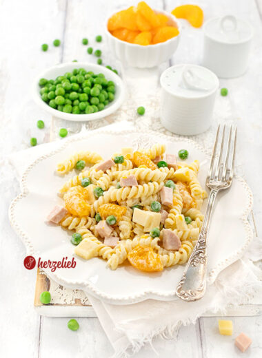 Nahaufnahme eines Pastasalats nach einem Nudelsalat mit Mandarinen Rezept von Oma auf einem weißen Teller, ergänzt durch grüne Erbsen und Würfel aus Schinken und Käse, präsentiert als frisches Rezept mit einer kleinen Schale voller Mandarinenstückchen im Hintergrund