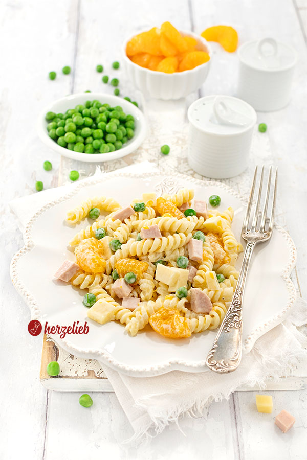 Nahaufnahme eines Pastasalats nach einem Nudelsalat mit Mandarinen Rezept von Oma auf einem weißen Teller, ergänzt durch grüne Erbsen und Würfel aus Schinken und Käse, präsentiert als frisches Rezept mit einer kleinen Schale voller Mandarinenstückchen im Hintergrund