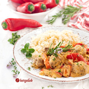 Teller mit einer Gemüsepfanne nach einem Paprikapfanne Rezept von herzelieb. In dem Pfannengericht sieht man kleine Fleischbällchen, rote Paprikastücke in cremiger Sauce. Serviert mit Reis, garniert mit frischen Kräutern. Im Hintergrund liegen frische rote Paprika und Rosmarinzweige auf weißem Untergrund.