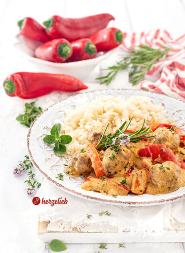 Teller mit einer Gemüsepfanne nach einem Paprikapfanne Rezept von herzelieb. In dem Pfannengericht sieht man kleine Fleischbällchen, rote Paprikastücke in cremiger Sauce. Serviert mit Reis, garniert mit frischen Kräutern. Im Hintergrund liegen frische rote Paprika und Rosmarinzweige auf weißem Untergrund.