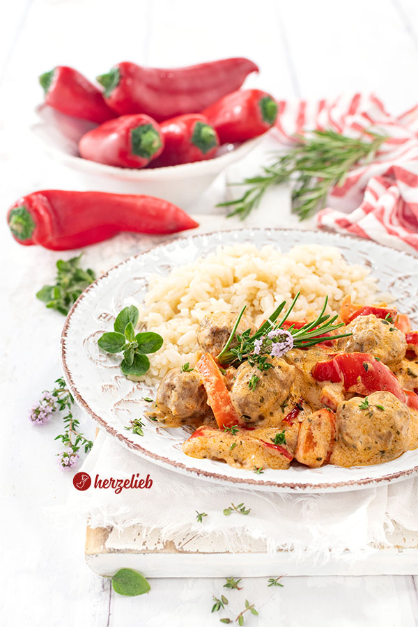 Teller mit einer Gemüsepfanne nach einem Paprikapfanne Rezept von herzelieb.  In dem Pfannengericht sieht man kleine Fleischbällchen, rote Paprikastücke in cremiger Sauce. Serviert mit Reis, garniert mit frischen Kräutern. Im Hintergrund liegen frische rote Paprika und Rosmarinzweige auf weißem Untergrund.