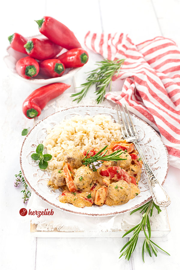 Gemüsepfanne kleinen Fleischbällchen, roten Paprikastücken und cremiger Sauce, serviert mit Reis nach einem Paprikapfanne Rezept von herzelieb auf einem Teller. Im Hintergrund frische rote Paprika in einer Schüssel und frische Kräuter auf weißem Holztisch. Das Pfannengericht ist bestreut mit Kräutern.