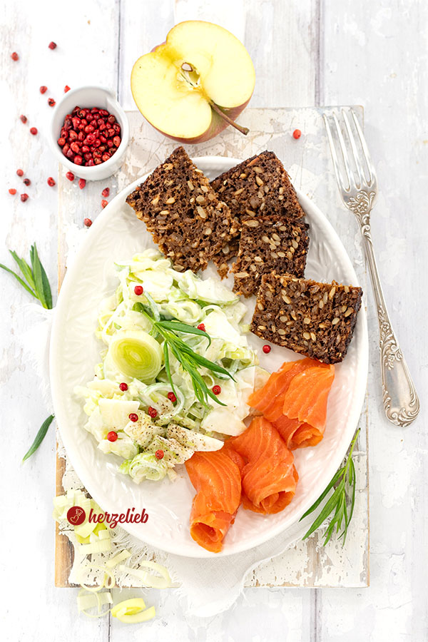 Ein Teller mit einem Lauch-Apfel-Salat nach einem Porre-Æblesalat Rezept aus Dänemark von herzelieb. Angerichtet als Beilage zum geräuchertem Lachs und Roggenbrot. Bestreut mit roten Pfefferbeeren, dekoriert mit Estragon. 