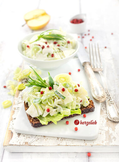Eine Scheibe Roggenbrot, belegt mit einem Salat nach einem Porre-Æblesalat Rezept von herzelieb. Bestreut mit roten Pfefferbeeren und dekoriert mit Estragon. Im Hintergrund eine Schüssel mit dem Lauchsalat, Besteck und ein halber Apfel.