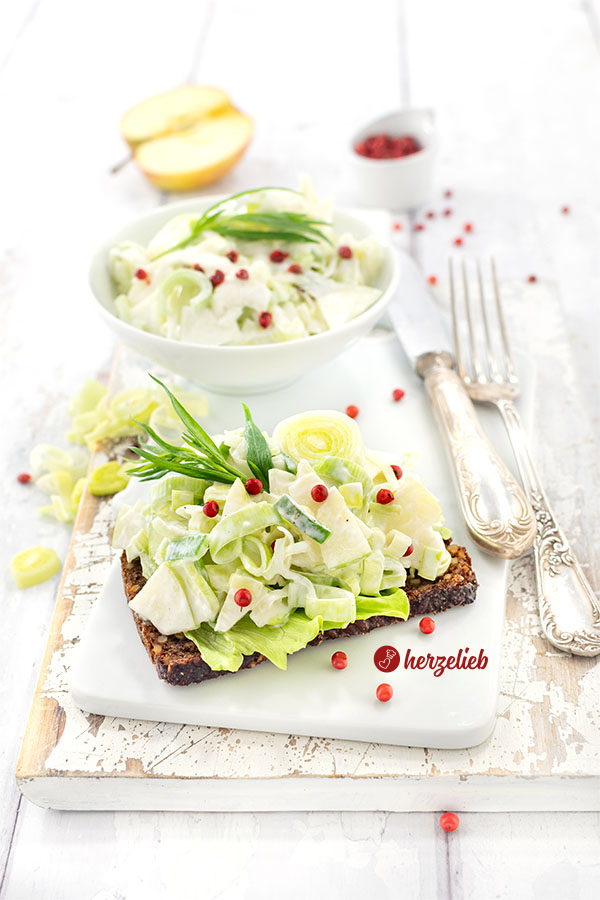 Eine Scheibe Roggenbrot, belegt mit einem Salat nach einem Porre-Æblesalat Rezept von herzelieb. Bestreut mit roten Pfefferbeeren und dekoriert mit Estragon. Im Hintergrund eine Schüssel mit dem Lauchsalat, Besteck und ein halber Apfel.