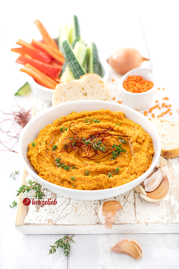Weiße Schale mit würzigem, rötlichem Linsendip nach einem Rote-Linsen-Feta-Dip Rezept von herzelieb.  Verfeinert und dekoriert mit Thymian und Chilifäden, begleitet von Paprika- und Gurkensticks sowie Brotscheiben.