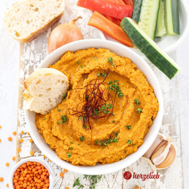 Von oben fotografiert: Rötlicher Fetadip mit Linsen nach einem Rote Linsen Feta Dip Rezept von herzelieb. Dekoriert mit Thymianzweigen und Chilifäden, daneben frisches Brot, rote Linsen, Zwiebel und Knoblauch. Im Hintergrund Gemüsesticks aus Gurke und Paprika