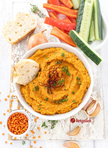 Von oben fotografiert: Rötlicher Fetadip mit Linsen nach einem Rote Linsen Feta Dip Rezept von herzelieb. Dekoriert mit Thymianzweigen und Chilifäden, daneben frisches Brot, rote Linsen, Zwiebel und Knoblauch. Im Hintergrund Gemüsesticks aus Gurke und Paprika