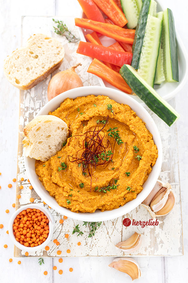 Von oben fotografiert: Rötlicher Fetadip mit Linsen nach einem Rote-Linsen-Feta-Dip Rezept von herzelieb. Dekoriert mit Thymianzweigen und Chilifäden, daneben frisches Brot, rote Linsen, Zwiebel und Knoblauch. Im Hintergrund Gemüsesticks aus Gurke und Paprika