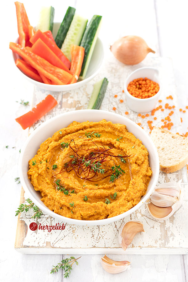 Schüssel mit cremigem Dip aus roten Linsen nach einem Rote-Linsen-Feta-Dip Rezept von herzelieb, garniert mit frischem Thymian und Chilifäden, serviert mit Brotscheiben sowie Gemüsesticks aus Gurke und Paprika