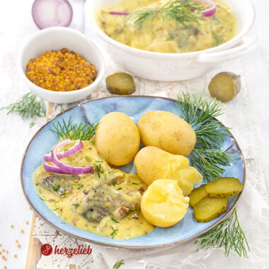Ein Teller mit Sennepssild aus Dänemark nach eiem Senfhering Rezept von herzelieb. Angerichtet mit Pellkartoffeln, Dill, Gurke, roten Zwiebeln Im Hintergrund eine Schale Senf und eine Schale mit dem eingelegtem Hering.
