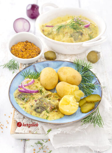 Ein Teller mit Sennepssild aus Dänemark nach eiem Senfhering Rezept von herzelieb. Angerichtet mit Pellkartoffeln, Dill, Gurke, roten Zwiebeln Im Hintergrund eine Schale Senf und eine Schale mit dem eingelegtem Hering.