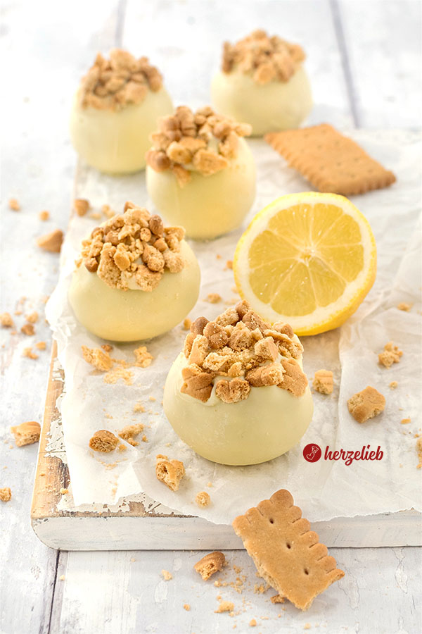 5 Zitronen-Käsekuchenkugeln auf einem weißen Brett. Umhüllt mit weißer Schokolade, bestreut mit Kekskrümeln. Im Hintergrund eine Zitrone und Butterkekse.