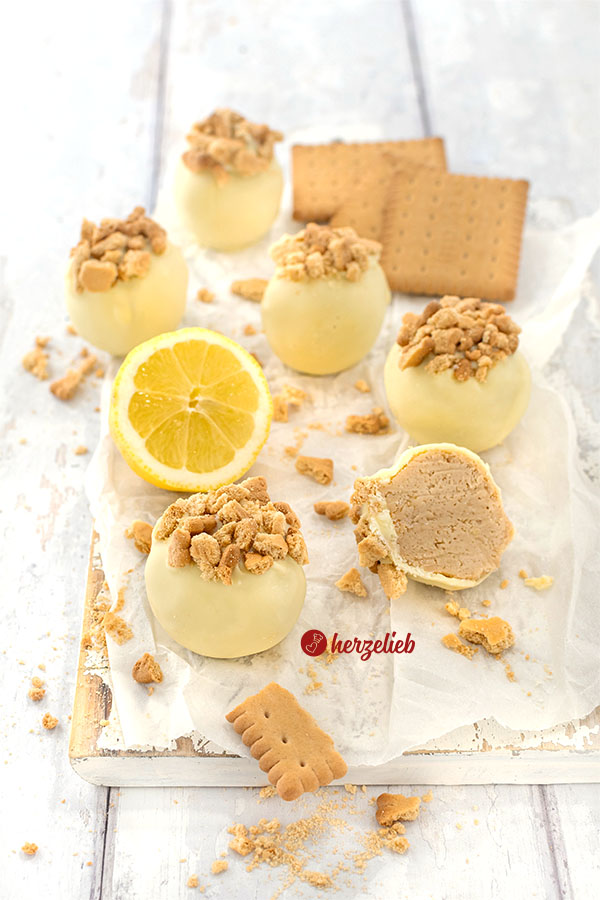 Einige Zitronen-Käsekuchenkugeln, die mit weißer Schokolade umhüllt und mit Kekskrümeln bestreut sind. Zwischen den Kugeln liegt eine halbe Zitrone. Im Hintergrund einige Butterkekse.