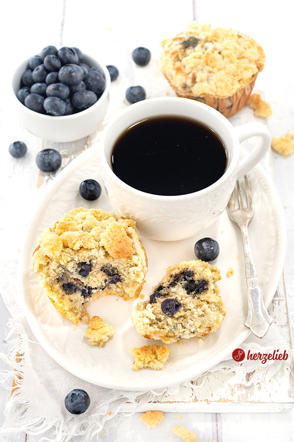 Ein saftiger Muffins mit Blaubeeren nach einem Blaubeermuffins Rezept von herzelieb. Der Muffins ist auf einem Teller angerichtet, daneben liegt eine Kuchengabel und dahinter eine Tasse Kaffee. Im Hintergrund noch ein Muffin und eine Schale mit Heidelbeeren.