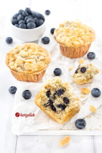 Drei Muffins mit Blaubeeren mit Streuseln nach einem Blaubeermuffins Rezept von herzelieb. Im Hintergrund eine Schale Blaubeeren. Zwischen den Kuchen verstreut liegen weitere Heidelbeeren.