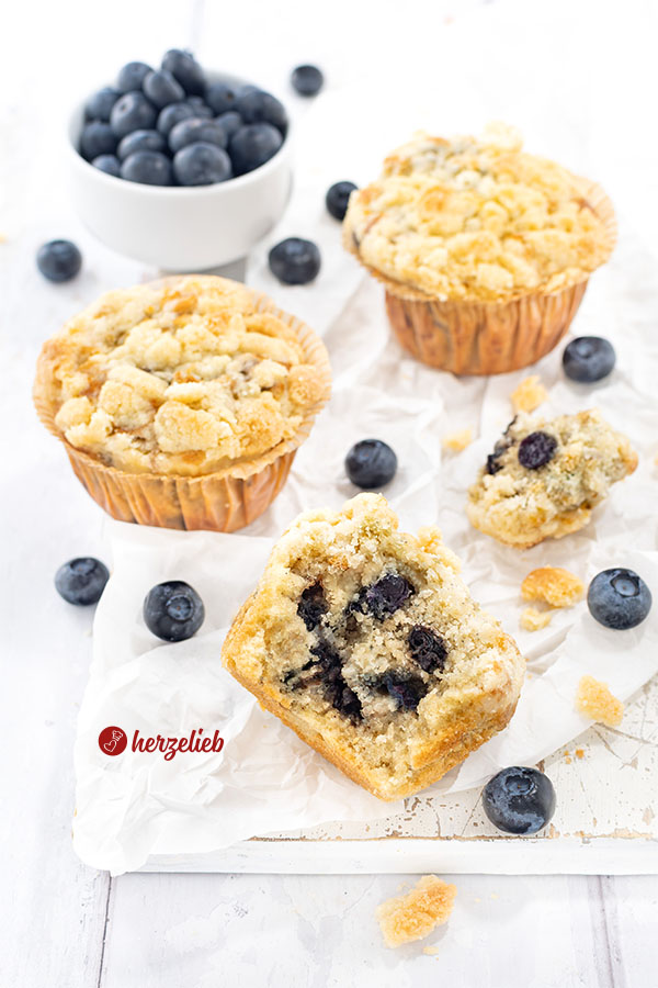 Drei Muffins mit Blaubeeren mit Streuseln nach einem Blaubeermuffins Rezept von herzelieb. Im Hintergrund eine Schale Blaubeeren. Zwischen den Kuchen verstreut liegen weitere Heidelbeeren.
