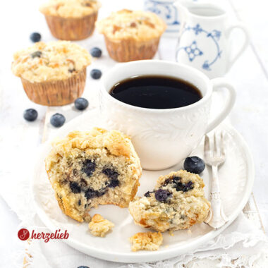 Ein Muffin mit Blaubeeren und Butterstreuseln nach einem Blaubeermuffins Rezept von herzelieb, serviert auf einem weißen Teller neben einer Tasse schwarzem Kaffee Im Hintergrund 3 weitere Muffins und eine Schale voller frischer Blaubeeren.