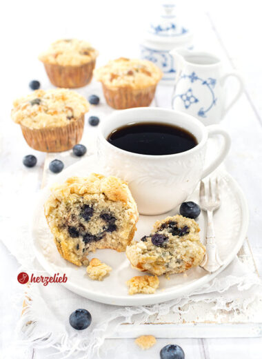 Ein Muffin mit Blaubeeren und Butterstreuseln nach einem Blaubeermuffins Rezept von herzelieb, serviert auf einem weißen Teller neben einer Tasse schwarzem Kaffee Im Hintergrund 3 weitere Muffins und eine Schale voller frischer Blaubeeren.