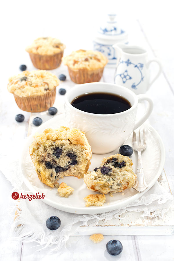 Ein Muffin mit Blaubeeren und Butterstreuseln nach einem Blaubeermuffins Rezept von herzelieb, serviert auf einem weißen Teller neben einer Tasse schwarzem Kaffee Im Hintergrund 3 weitere Muffins und eine Schale voller frischer Blaubeeren.