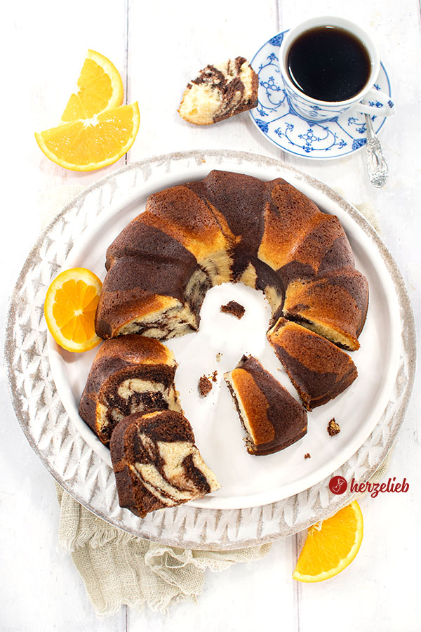 Ein angeschnittener Orangen-Marmorkuchen in Gugelhupf-Form auf einem weißen Teller, umgeben von frischen Orangenscheiben und einer Tasse schwarzem Kaffee in blauweißen Porzellan. 