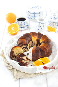 Der Gugelhupf-Orangen-Marmorkuchen ist frisch angeschnitten in einer weißen Form zu sehen, mit Orangenscheiben daneben und einer Tasse Kaffee in blau-weißem Geschirr.