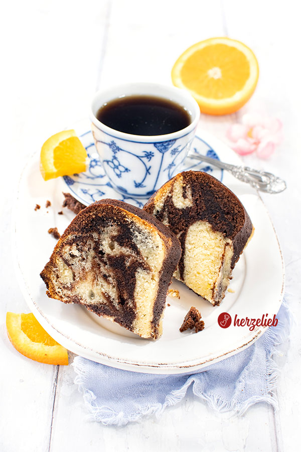 Zwei Scheiben des saftigen Orangen-Marmorkuchens liegen auf einem kleinen Teller neben einer Tasse Kaffee, dekoriert mit Orangenscheiben und einer halbierten Orange im Hintergrund.