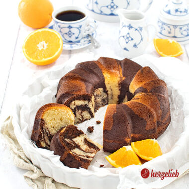 Der Gugelhupf-Orangen-Marmorkuchen ist frisch angeschnitten in einer weißen Form zu sehen, mit Orangenscheiben daneben und einer Tasse Kaffee in blau-weißem Geschirr.
