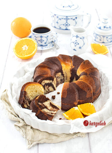 Der Gugelhupf-Orangen-Marmorkuchen ist frisch angeschnitten in einer weißen Form zu sehen, mit Orangenscheiben daneben und einer Tasse Kaffee in blau-weißem Geschirr.