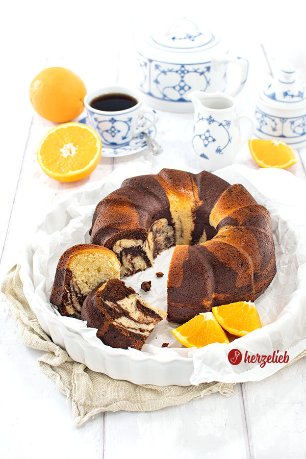 Der Gugelhupf-Orangen-Marmorkuchen ist frisch angeschnitten in einer weißen Form zu sehen, mit Orangenscheiben daneben und einer Tasse Kaffee in blau-weißem Geschirr.