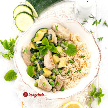 Eine weiße Schale mit einem Puten-Zucchini-Topf, bestehend aus gebratenem Putengeschnetzeltem, grünen Erbsen und gewürfelter Zucchini in einer hellen Käsesoße, garniert mit frischer Minze. Dazu wird Reis serviert. Im Hintergrund liegen frische Zucchinischeiben, ein ganzes Stück Zucchini, ein Salzstreuer, Zitronenscheiben und weitere frische Kräuter auf einem hellen Untergrund