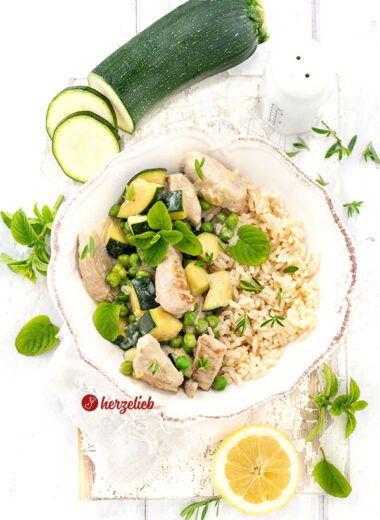 Eine weiße Schale mit einem Puten-Zucchini-Topf, bestehend aus gebratenem Putengeschnetzeltem, grünen Erbsen und gewürfelter Zucchini in einer hellen Käsesoße, garniert mit frischer Minze. Dazu wird Reis serviert. Im Hintergrund liegen frische Zucchinischeiben, ein ganzes Stück Zucchini, ein Salzstreuer, Zitronenscheiben und weitere frische Kräuter auf einem hellen Untergrund
