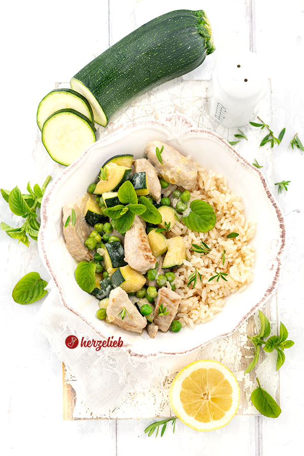 Eine weiße Schale mit einem Puten-Zucchini-Topf, bestehend aus gebratenem Putengeschnetzeltem, grünen Erbsen und gewürfelter Zucchini in einer hellen Käsesoße, garniert mit frischer Minze. Dazu wird Reis serviert. Im Hintergrund liegen frische Zucchinischeiben, ein ganzes Stück Zucchini, ein Salzstreuer, Zitronenscheiben und weitere frische Kräuter auf einem hellen Untergrund