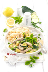 Ein Teller mit Puten-Zucchini-Topf in cremiger Käsesoße, garniert mit frischer Minze und grünen Erbsen, dazu eine Portion Reis. Im Hintergrund liegen frische Zitronen, Zucchinischeiben sowie Salz- und Pfefferstreuer auf einem weißen Holztisch. Ein silberner Tellergabel liegt rechts auf dem Teller.