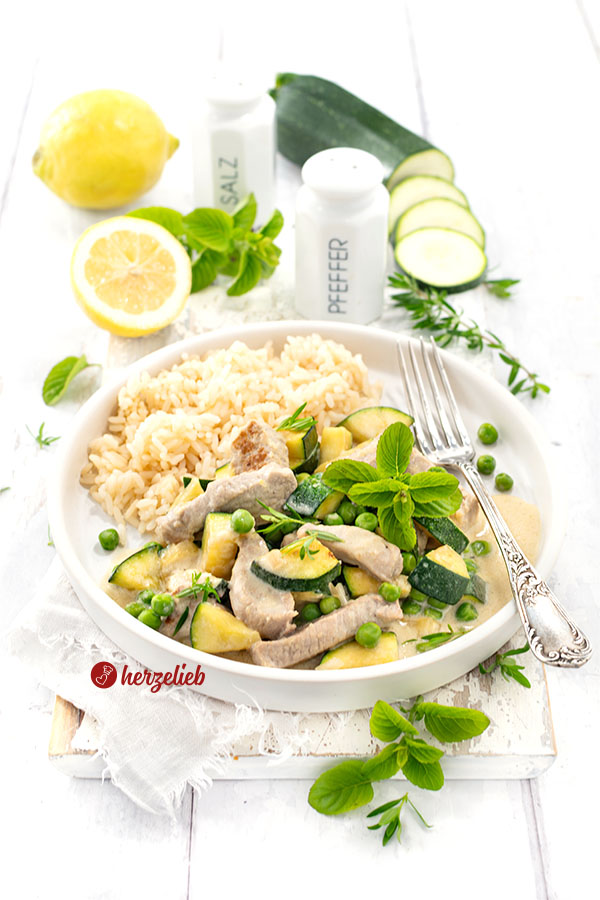 Ein Teller mit Puten-Zucchini-Topf in cremiger Käsesoße, garniert mit frischer Minze und grünen Erbsen, dazu eine Portion Reis. Im Hintergrund liegen frische Zitronen, Zucchinischeiben sowie Salz- und Pfefferstreuer auf einem weißen Holztisch. Ein silberner Tellergabel liegt rechts auf dem Teller.