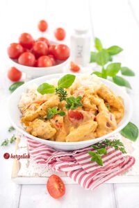 Ein Teller mit Putengeschnetzeltes in cremiger Tomaten-Frischkäse-Soße, serviert mit Reis und garniert mit frischen Kräutern, zeigt das fertige Gericht dieses beliebten Rezeptes. Im Hintergrund sind frische Tomaten in einer Schale zu sehen.